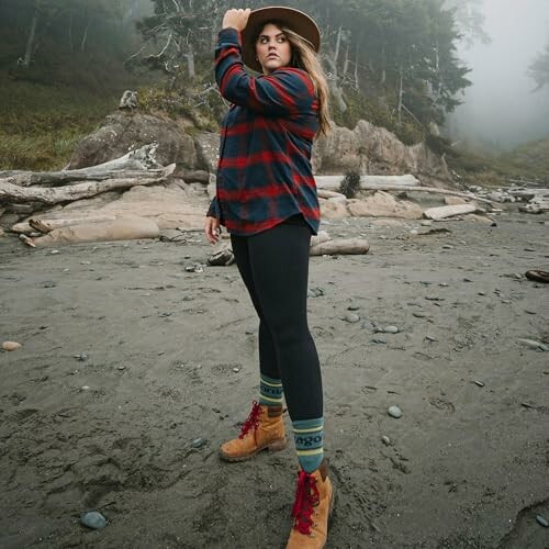 Woman standing on a foggy beach wearing a hat and plaid shirt.